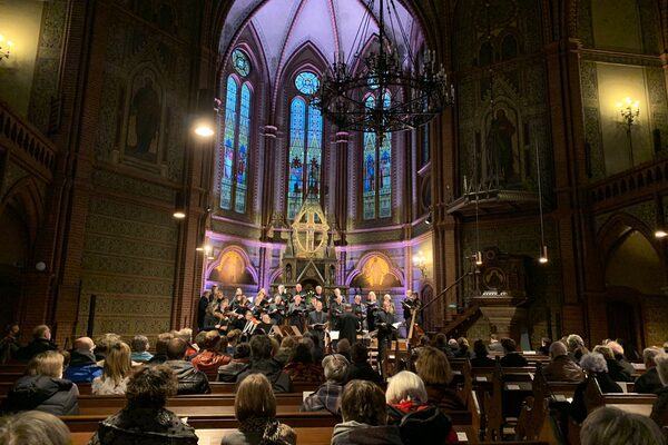 Der Chor in befüllter Kirche. Sie stehen vor einer großen Rundung, die Violett beleuchtet ist. Alles sieht sehr edel aus.