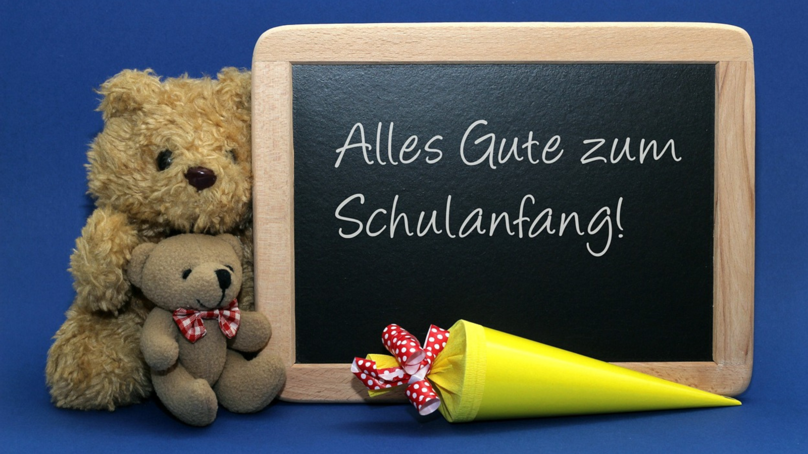 Blackboard with text: all the best for starting school. You can see two stuffed bears to the left of the blackboard and a yellow school cone in the foreground