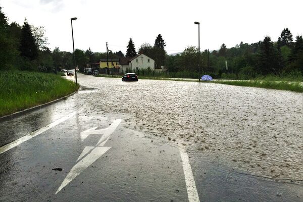 Eine überflutete Straße im Regen, im Wasser auf der Straße sind zwei Autos zu sehen, ein lila Regenschirm liegt im Hintergrund am Straßenrand.