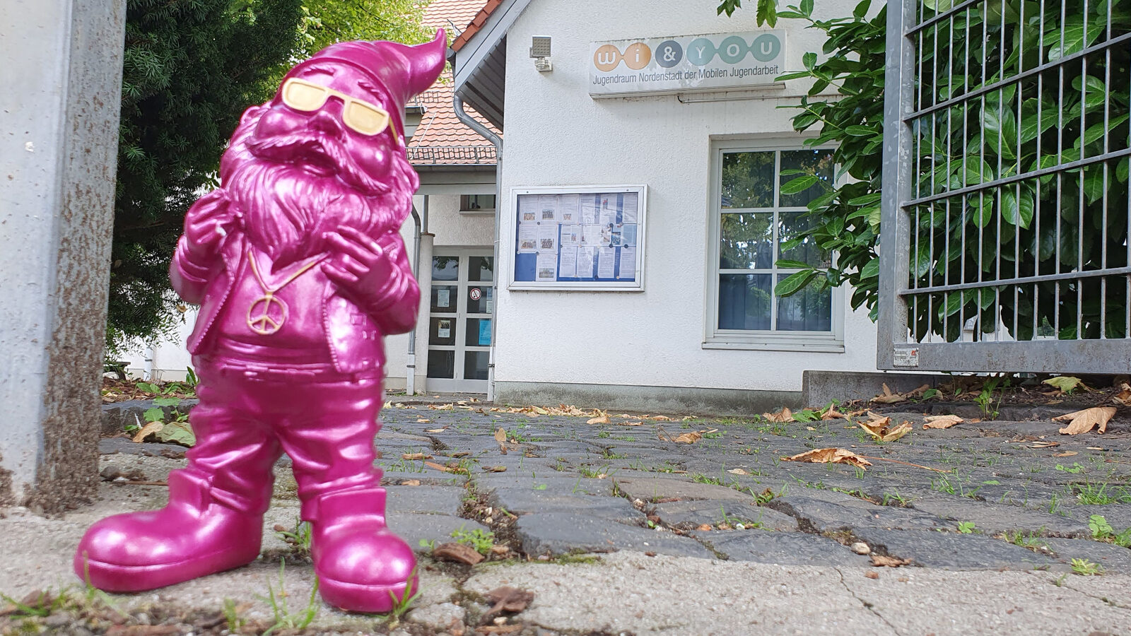 Ein pinkfarbener Zwerg steht auf dem Pflaster. Dahinter ist ein weißes Haus mit einem Aushangkasten und einem Schild "Wi&You Jugendraum Nordenstadt Mobile Jugendarbeit".